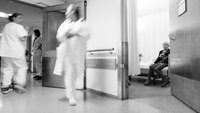 Bildet viser pasient i senga som venter på hjelp fra opptatt personale som fyker forbi.